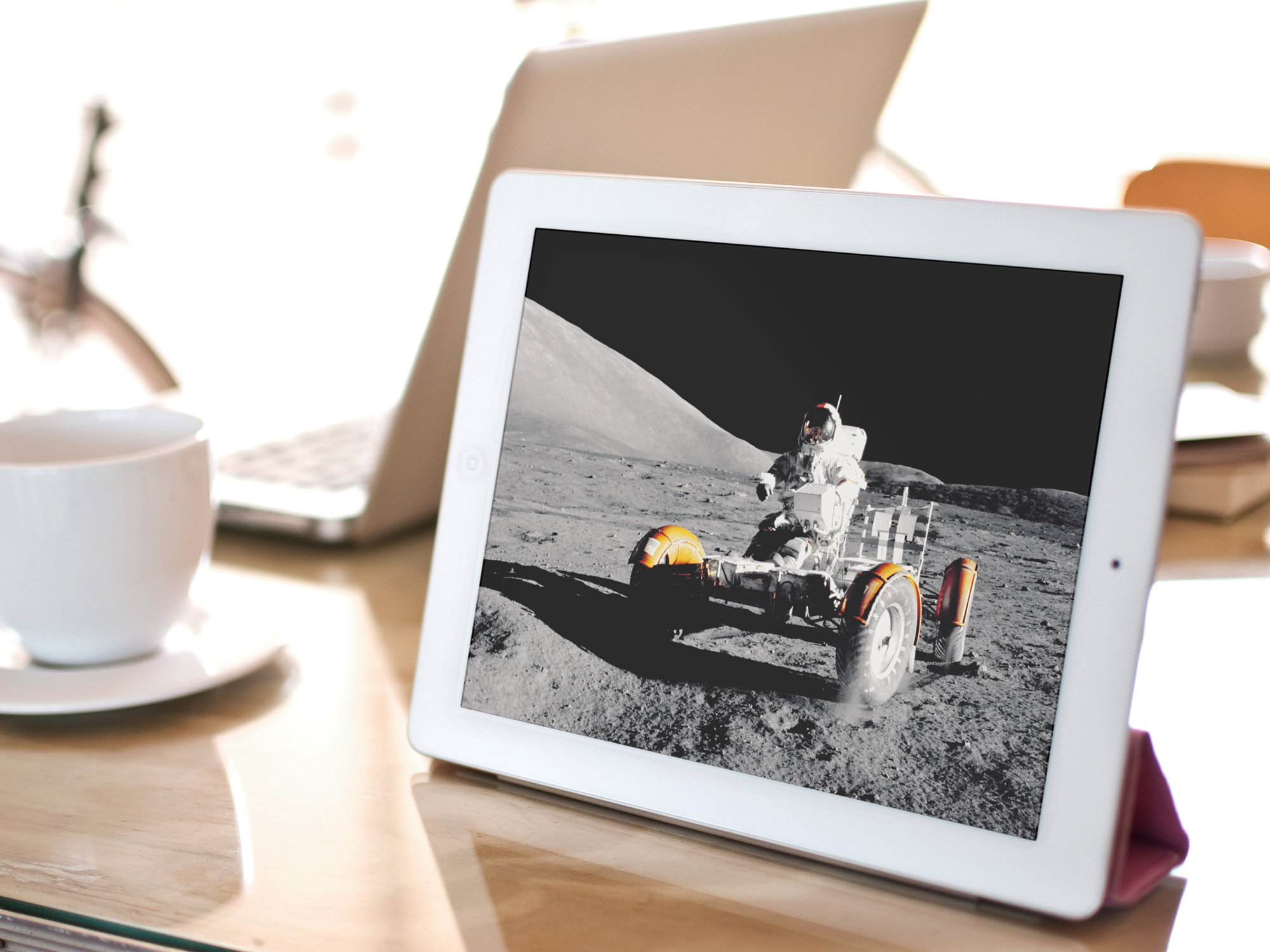 An ipad is propped up on a table. To the left is a coffee cup and in the background is an open laptop.