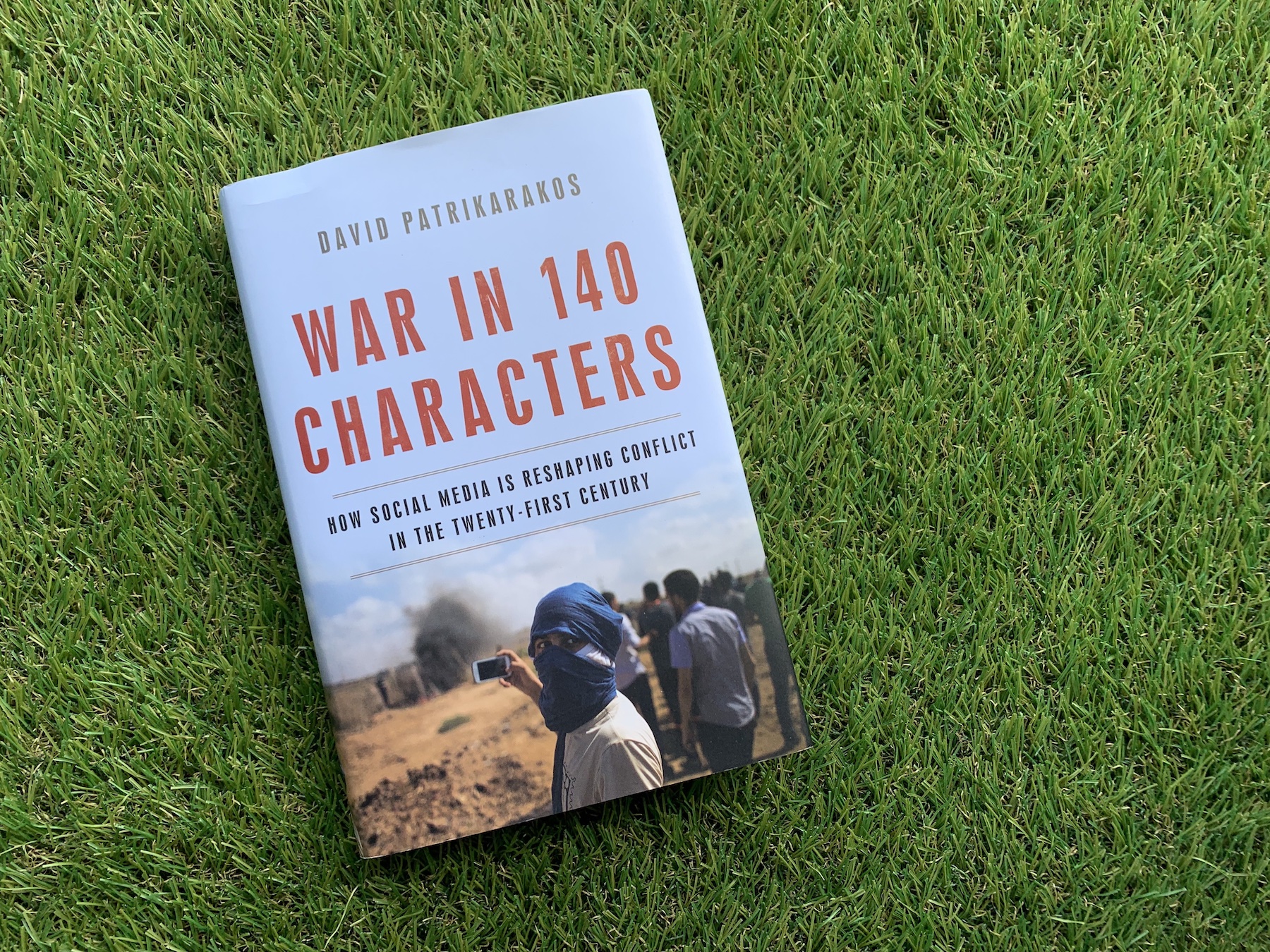 A photo of the book "War in 140 characters" by David Patrikarakos is on top of a rug that looks like green grass.